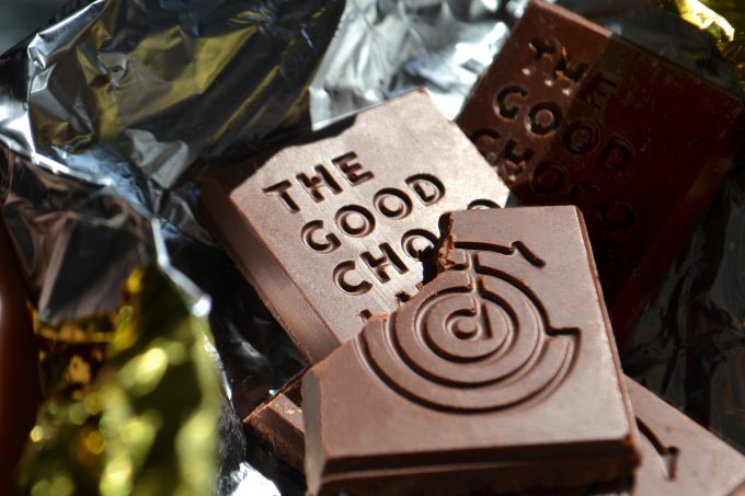 Close-up of a piece of The Good Chocolate bar with an embossed logo design, resting on an opened foil wrapper. One corner of the chocolate piece has a bite taken out of it, revealing a rich, smooth texture.