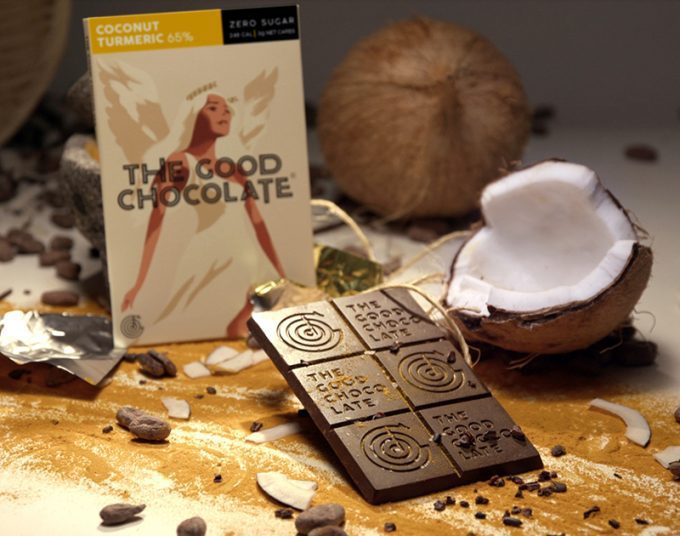 A package of The Good Chocolate's Coconut Turmeric 65% Zero Sugar dark chocolate bar is displayed alongside an unwrapped chocolate bar, pieces of coconut, and cocoa beans.