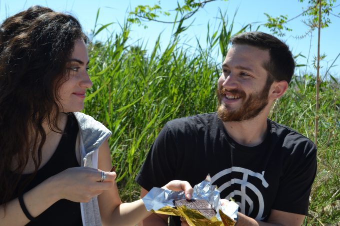 A man and a woman enjoy sharing a bar of "The Good Chocolate" outdoors on a sunny day, with greenery in the background. The man is smiling, and the woman is holding a piece of chocolate.