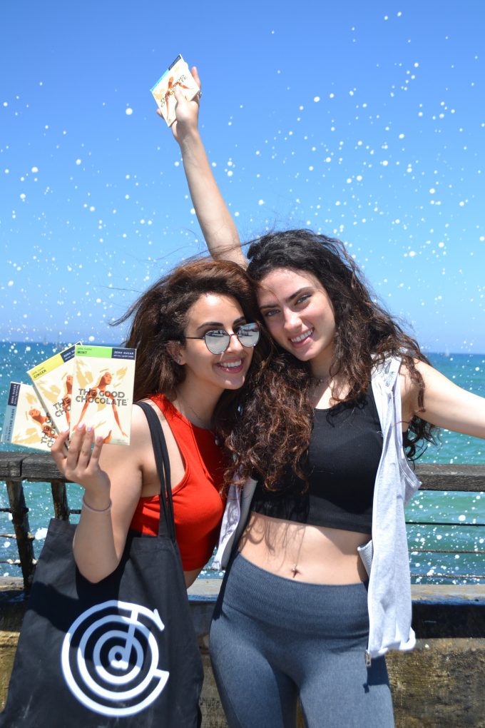 Two women smiling and holding The Good Chocolate bars by the seaside, one of them carrying a black tote bag with The Good Chocolate logo.