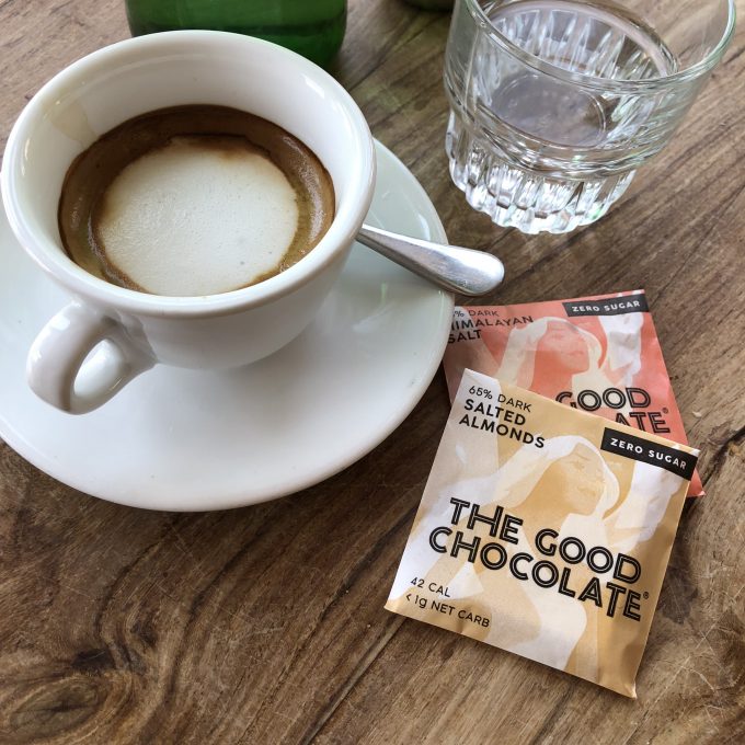 Two packs of The Good Chocolate's salted almond and Himalayan & sea salt squares , surrounded by a cup of coffee, next to a glass of water.