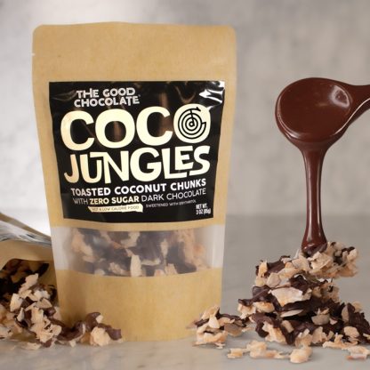 A package of The Good Chocolate's Coco Jungles toasted coconut chunks with zero sugar dark chocolate is displayed. Next to the package is a pile of the chocolate-coated coconut chunks, and a spoon dripping with melted dark chocolate.
