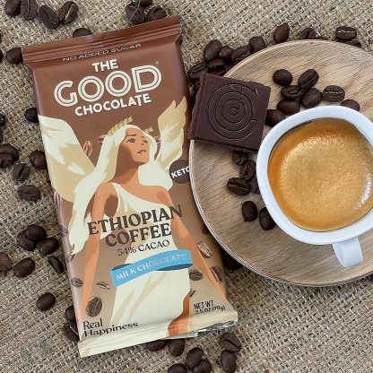 A package of The Good Chocolate Ethiopian Coffee dark chocolate bar, surrounded by coffee beans on a textured surface, next to a wooden plate with a square of chocolate and a cup of espresso.